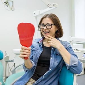 A middle-aged woman wearing glasses looks at her smile in the mirror while at the dentist’s office