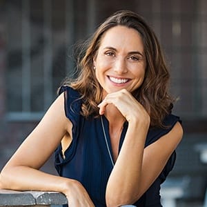 A woman wearing a navy blouse and sitting at a table outside smiles after seeing a trusted implant dentist