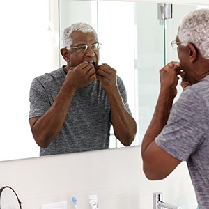 An older man flosses his teeth in the bathroom to remove bad bacteria from around his dental implants and natural teeth