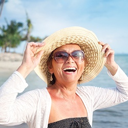 An older woman holding her hat while smiling on the beach after receiving implant dentures in Fairfax